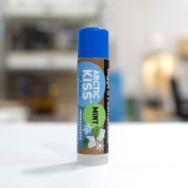 A single tube of B&amp;F lip balm standing upright on a white surface with a blurred indoor background. The product is Arctic Kiss Mint, featuring a blue cap, brown label, and blue, green, and white winter-themed design.