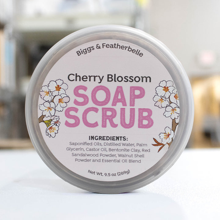 A round jar of Biggs &amp; Featherbelle Cherry Blossom Soap Scrub, featuring a white label with pink lettering and cherry blossom illustrations, set against a softly blurred background.