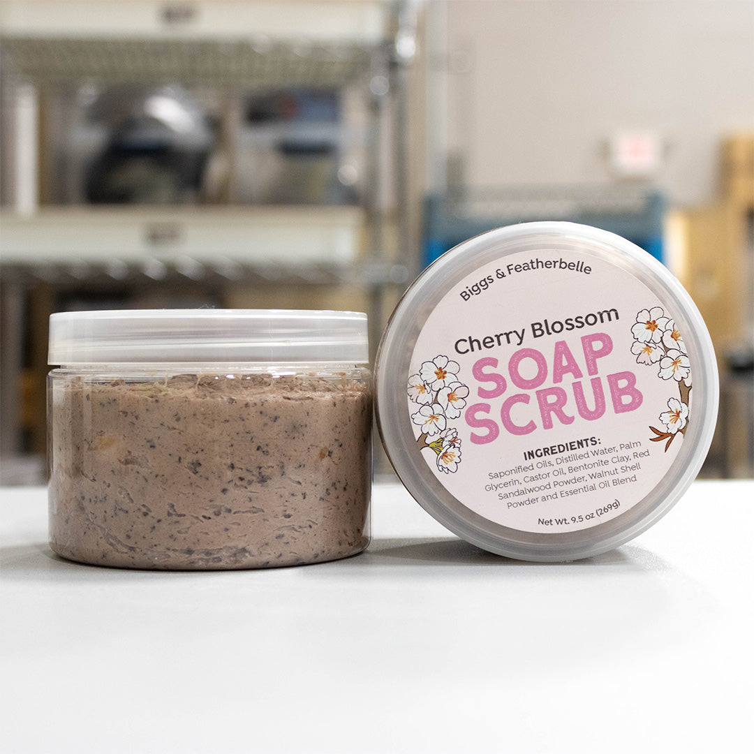 A jar of Biggs & Featherbelle Cherry Blossom Soap Scrub shown next to its lid on a white surface, with a softly blurred industrial background.