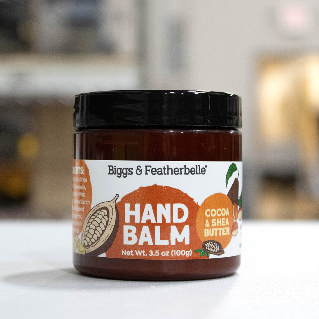 A jar of Biggs & Featherbelle' Hand Balm, Cocoa & Shea Butter, featuring a black lid and a white label with orange and brown graphics, set against a blurred background.