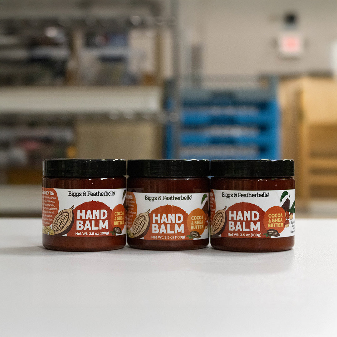Three jars of Biggs & Featherbelle Hand Balm, specifically Cocoa & Shea Butter, lined up on a white surface with a blurred warehouse or storage room background.