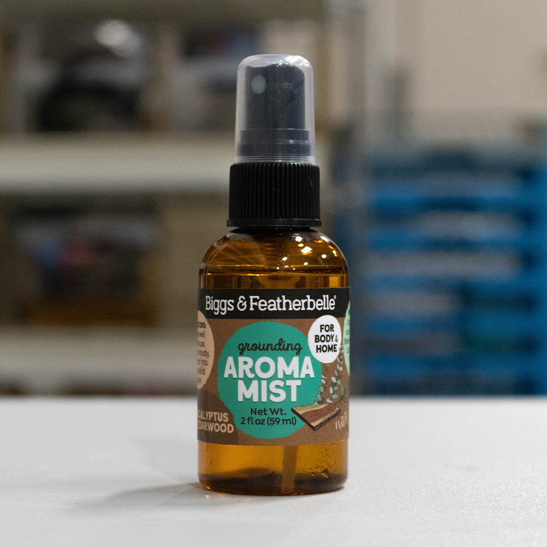 A 2 fl oz amber glass spray bottle of Biggs & Featherbelle Grounding Aroma Mist, featuring a black spray cap and a label with green and white graphics, set against a blurred indoor background.