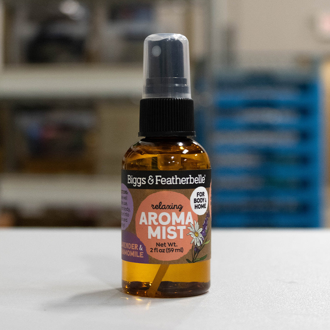 A bottle of Biggs & Featherbelli Relaxing Aroma Mist in lavender and chamomile, featuring an amber glass bottle, black spray cap, and a purple and orange label, set against a blurred indoor background.