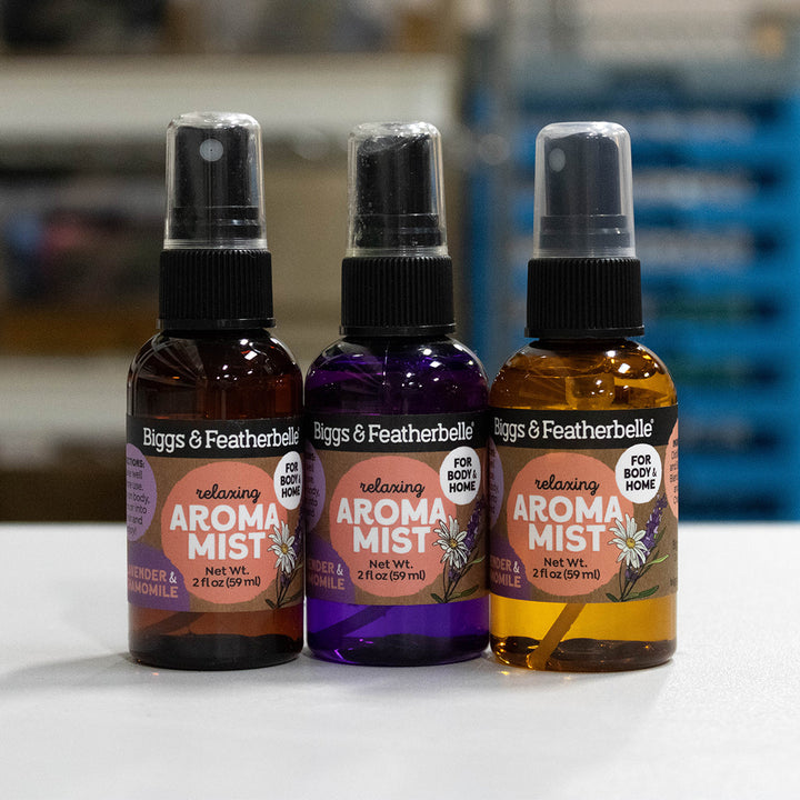 Three bottles of Biggs &amp; Featherbell relaxing Aroma Mist lined up on a white surface. The left bottle is amber with a black spray top, the middle bottle is purple with a black spray top, and the right bottle is golden yellow with a black spray top.