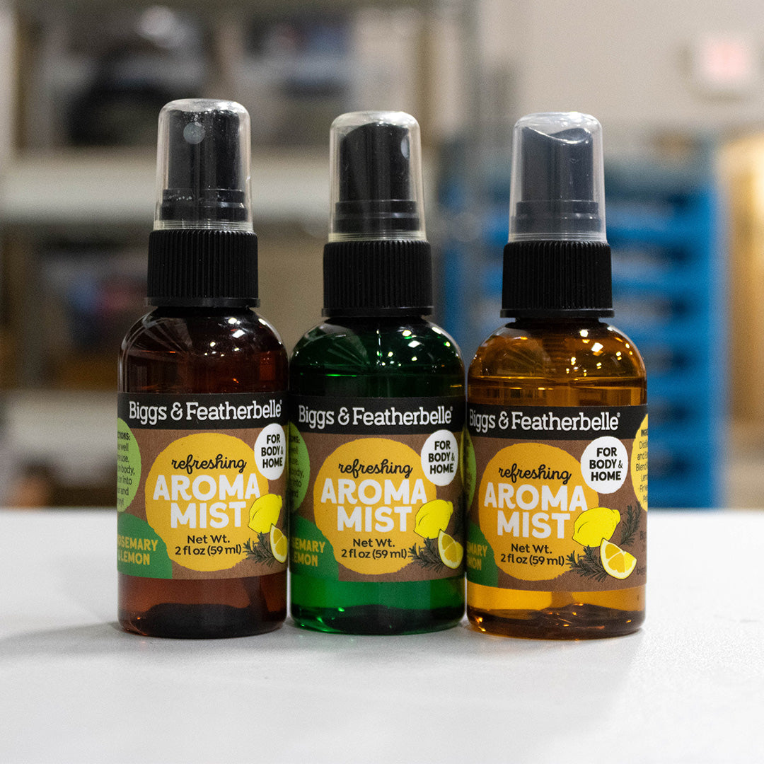 Three Biggs & Featherbelle refreshing Aroma Mist spray bottles lined up on a white surface with a blurred indoor background. The bottles are amber brown, green, and clear with black spray caps, featuring yellow and green labels.