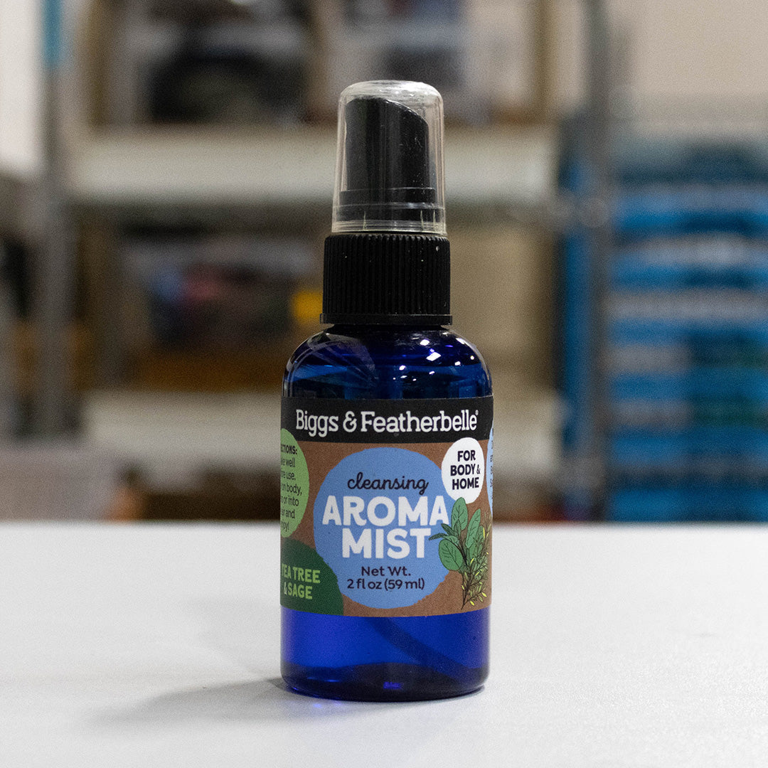 A 2 fl oz blue glass spray bottle of Biggs & Featherbelli Cleansing Aroma Mist, featuring a black spray cap and a colorful label, set against a blurred indoor background.