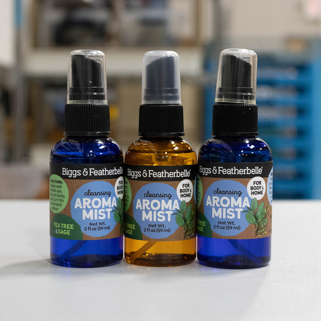 Three bottles of Biggs &amp; Featherbelle Cleansing Aromat Mist lined up on a white surface with a blurred indoor background. The left bottle is blue with a green label, the middle bottle is amber with a green label, and the right bottle is cobalt blue with a light blue label.