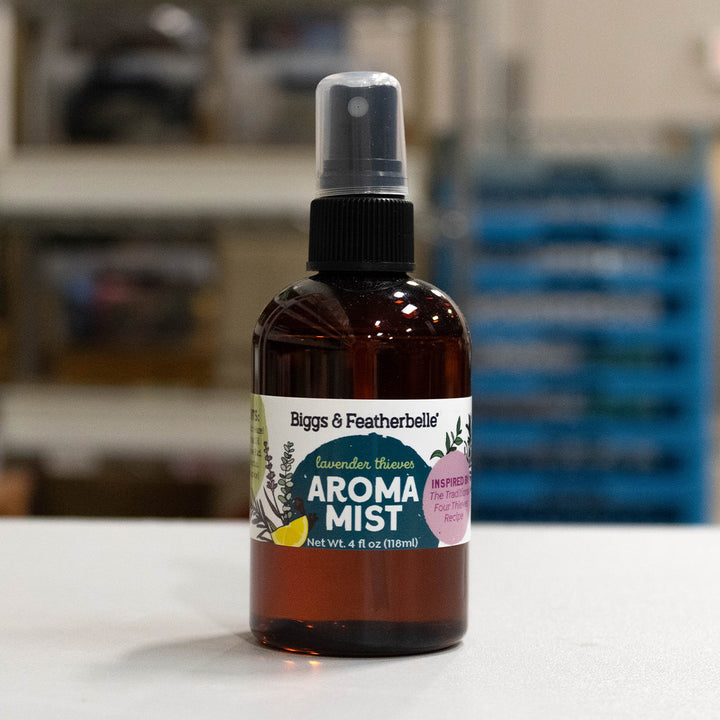A 4 fl oz amber glass spray bottle of Biggs &amp; Featherbelle Lavender Thieves Aroma Mist, standing on a white surface with a blurred indoor background.