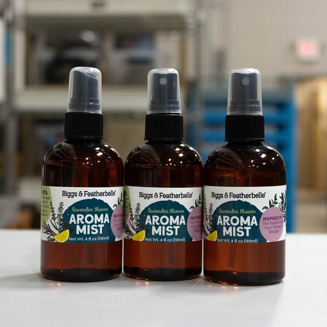 Three amber glass spray bottles of Biggs & Featherbelle Aroma Mist with black spray tops, lined up on a white surface with a blurred workshop or storage room background.