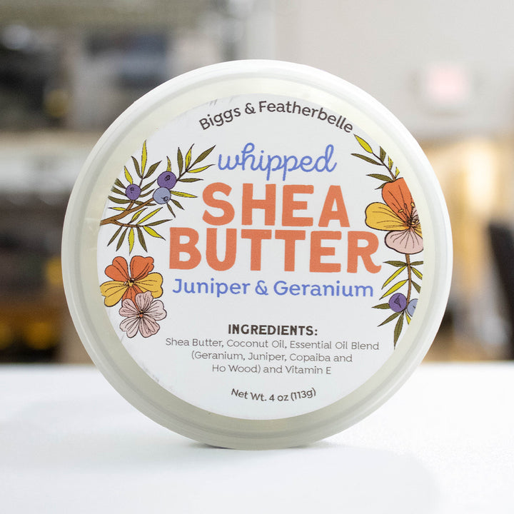 A round white tub of Biggs &amp; Featherbelle whipped Shea Butter, Juniper &amp; Geranium scent, shown from a top-down view on a white surface with a blurred background.