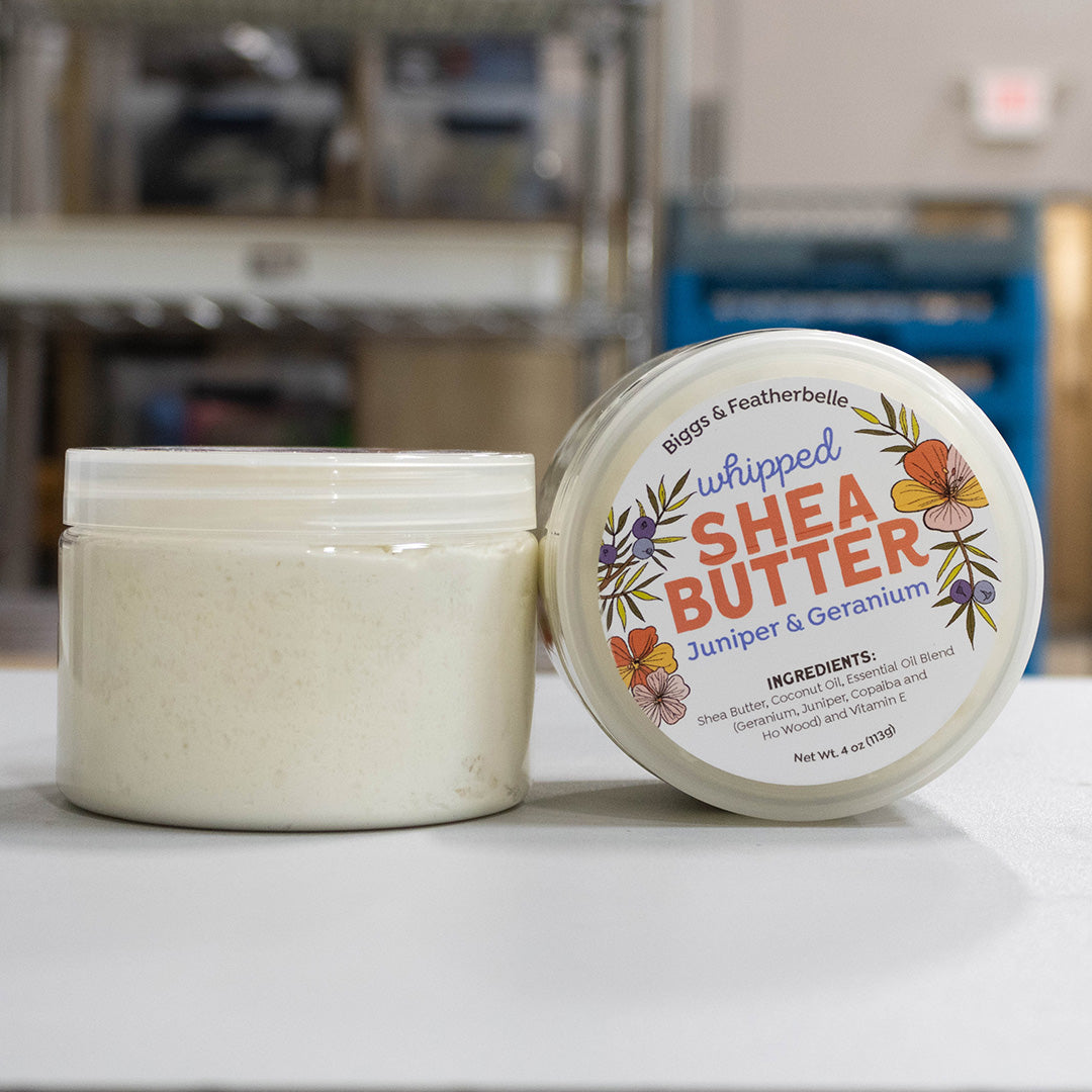 An open jar and matching lid of Biggs & Featherbelle Whipped Shea Butter on a white surface, with a softly blurred indoor background.