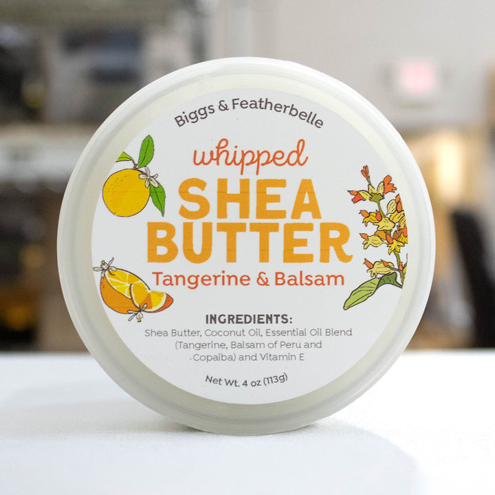 A round white plastic tub of Biggs &amp; Featherbelle Whipped Shea Butter in Tangerine &amp; Balsam, featuring a white lid with orange and yellow illustrations of tangerine and a balsam flower, set against a blurred background.