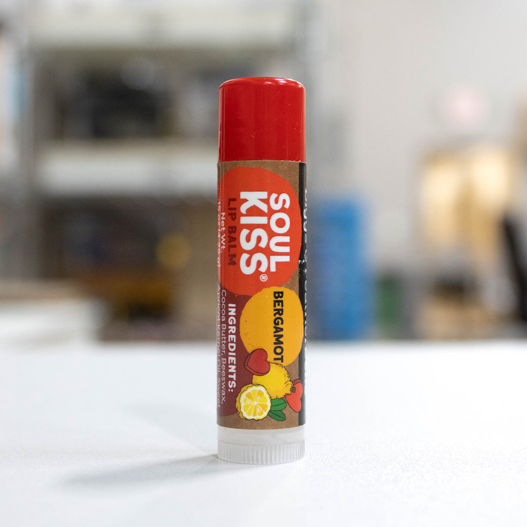 A single tube of B&F Soul Kiss Bergamot Lip Balm standing upright on a white surface with a blurred indoor background.