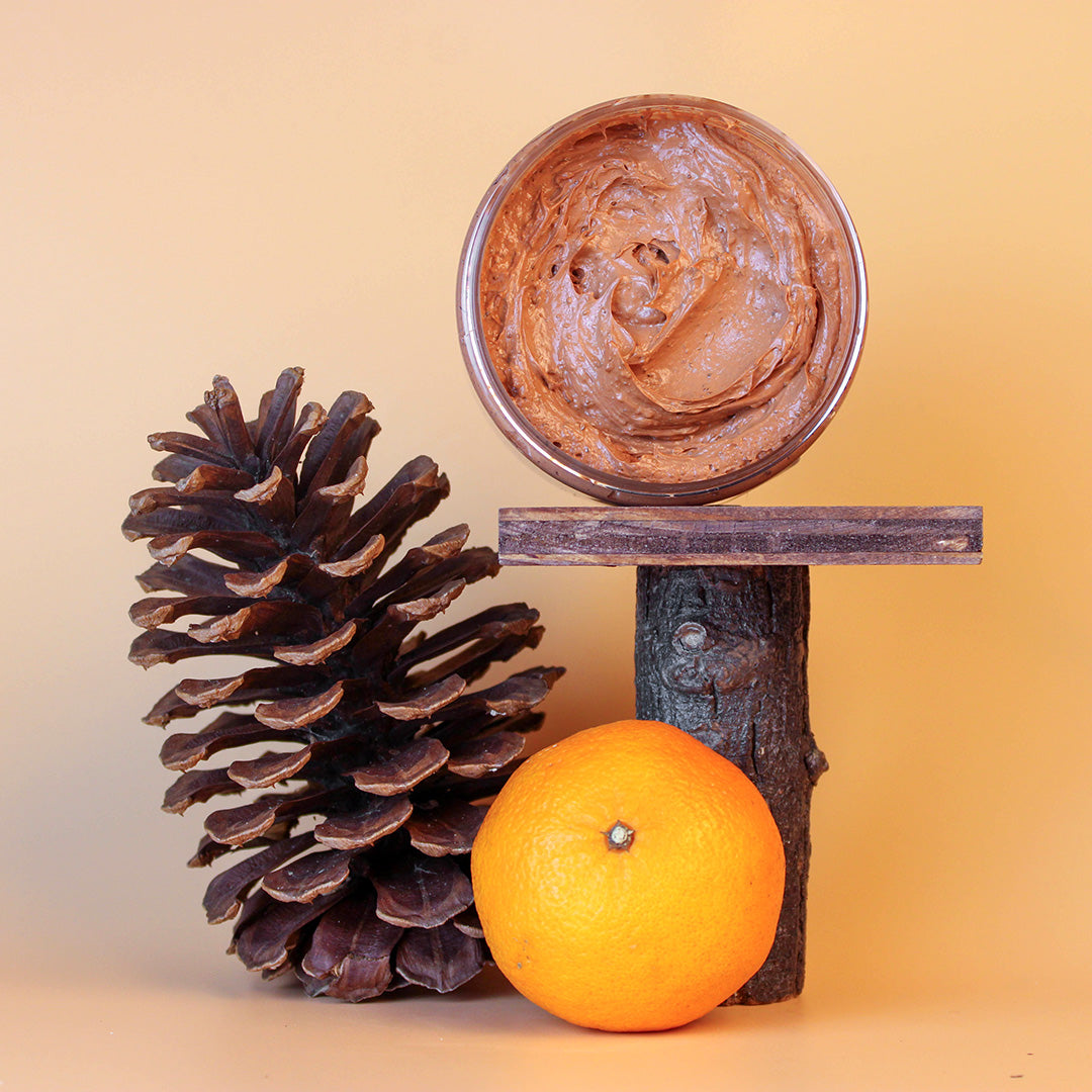 Pine cone, orange, and container of cedar soap scrub on a wooden stand against a beige background